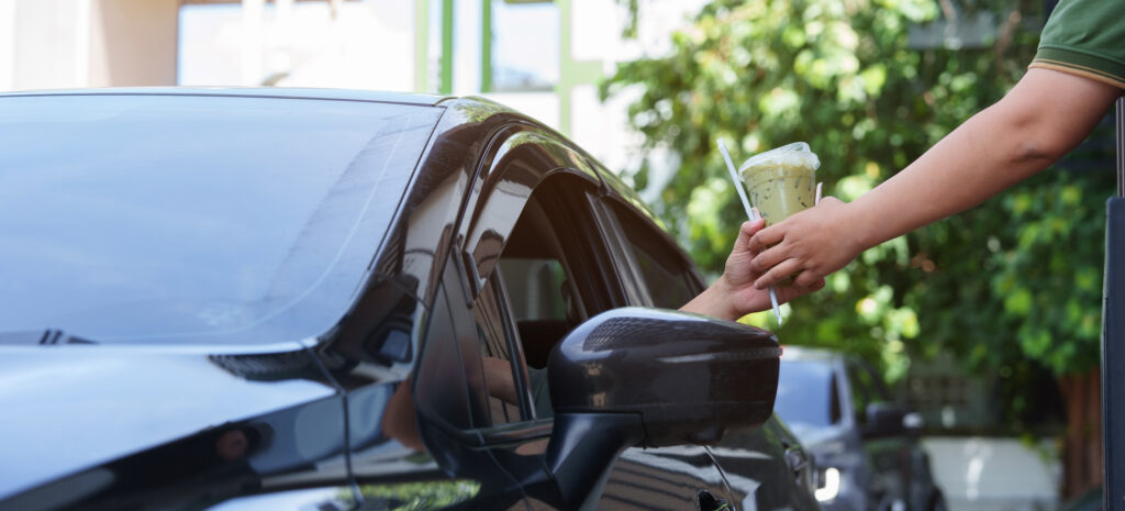 Person in a car receives an iced drink through the driver’s side window from someone outside, suggesting a drive-through or curbside pickup setting.