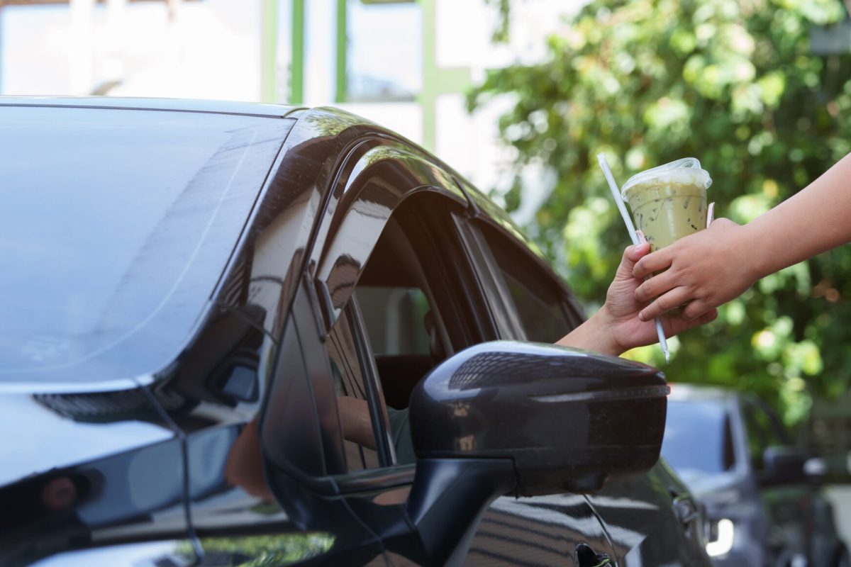 Person in a car receives an iced drink through the driver’s side window from someone outside, suggesting a drive-through or curbside pickup setting.